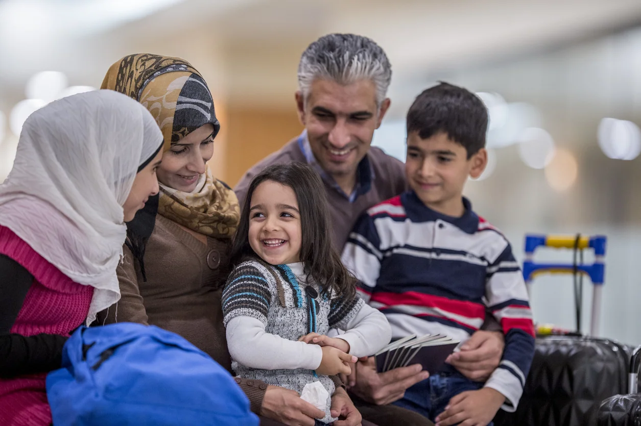 A Middle-eastern father, mother, brother and two sisters have just arrived to a new country. They are happily sitting together and smiling. The father is holding his family's passports. Their luggage is sitting to the side.